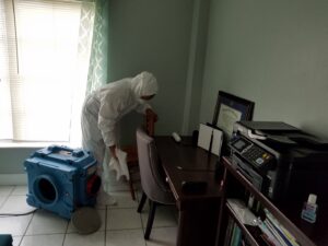 A technician in a protective suit cleaning a desk area with an air mover visible, indicating water damage restoration by Southcoast Cleaning Solutions & Restoration in Fall River, MA.