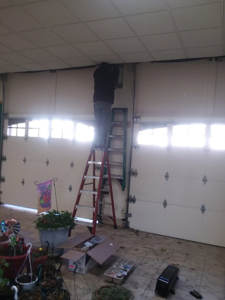 A technician on a ladder repairing the mechanism of a garage door inside a garage for PS GARAGE DOORS in Grand Forks, ND