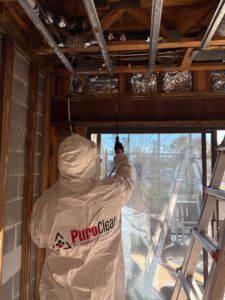 A PuroClean technician in a hazmat suit spraying a solution for mold remediation on exposed ceiling and wall framing in Bluffton, SC.