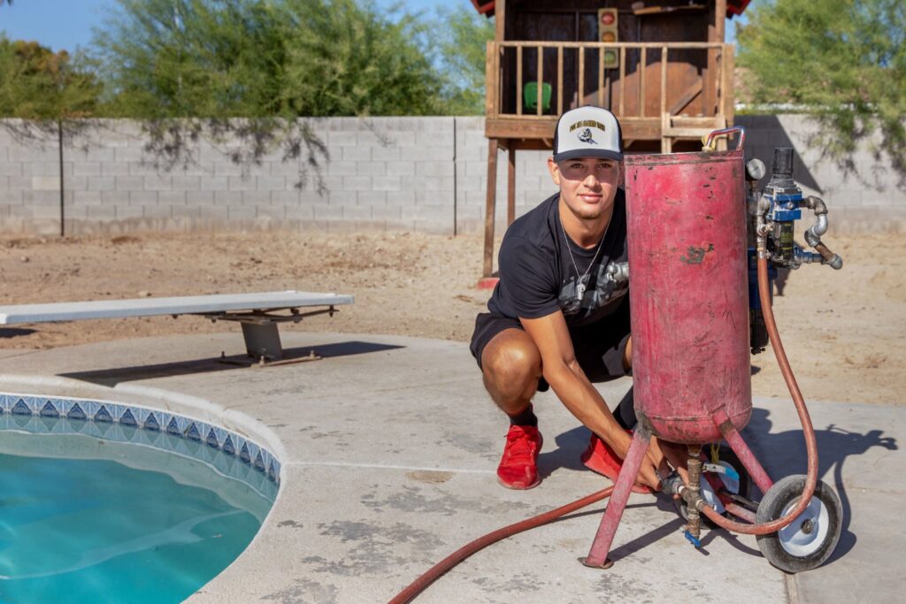 A technician from Pool Tile Cleaning Vegas crouching next to a pool cleaning blaster by a residential pool in Las Vegas, NV.