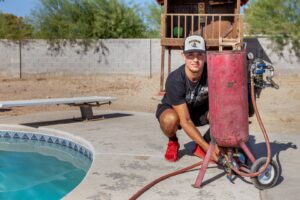 A technician from Pool Tile Cleaning Vegas crouching next to a pool cleaning blaster by a residential pool in Las Vegas, NV.