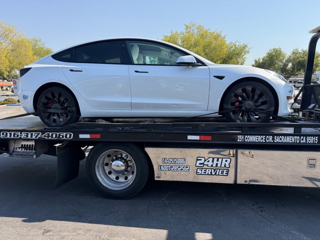 A white Tesla sedan being towed by an AJ's Towing Services & Roadside Assistance flatbed truck in Sacramento, CA.
