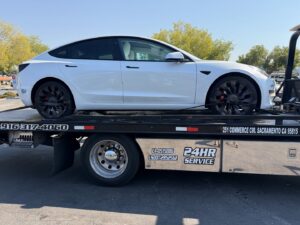 A white Tesla sedan being towed by an AJ's Towing Services & Roadside Assistance flatbed truck in Sacramento, CA.