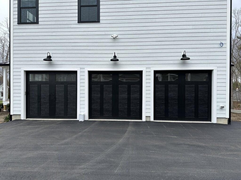 Three modern black garage doors with top windows on a light-colored house, installed by Steve Shumsky Unlimited Overhead Doors LLC in Hudson, NH