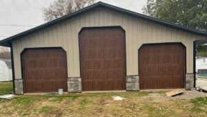 Three new brown garage doors with decorative stone pillars installed on a beige metal building by 4thgenerationsdoors in Huntington, WV.