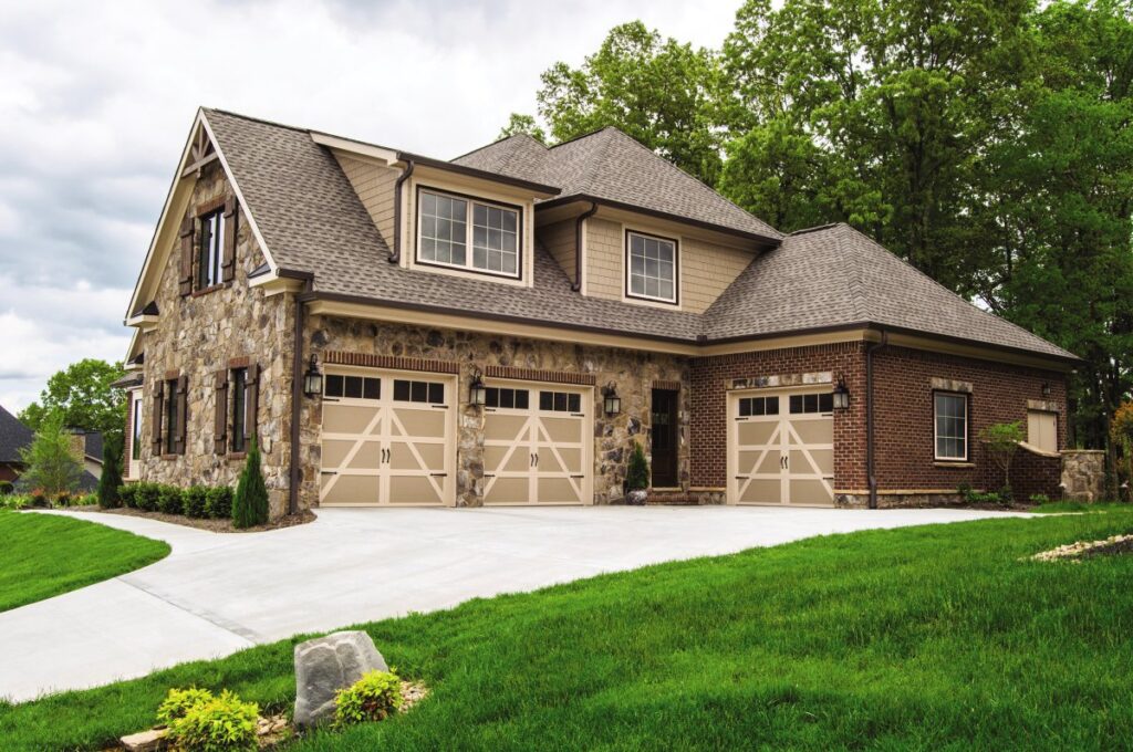 Three light-colored X-brace style garage doors on a stone and brick home by Access Door Systems in Uniontown, PA.