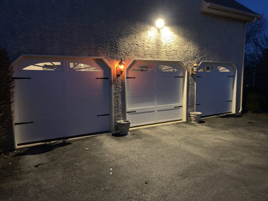 Three white carriage-style garage doors with decorative windows and hardware, illuminated at night by 4thgenerationsdoors in Huntington, WV.