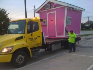 A Tow Jam flatbed tow truck transporting a small pink shed or building in Dallas, TX.