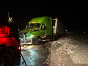 A tow truck from Select Towing and Recovery assisting a green semi-truck stuck in deep snow at night in Casper, WY.