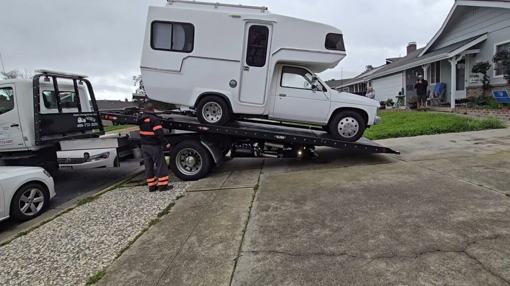A tow truck driver loading a white pickup truck with a camper onto a flatbed for Ibanez Towing Service LLC in San Jose, CA.
