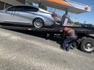 A tow truck driver securing a silver sedan onto a flatbed tow truck for Ibanez Towing Service LLC in San Jose, CA.