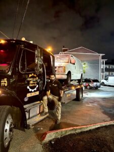 A tow truck driver from Safety AUTO Towing LLC in Seattle, WA, standing next to a flatbed truck towing a white van at night.