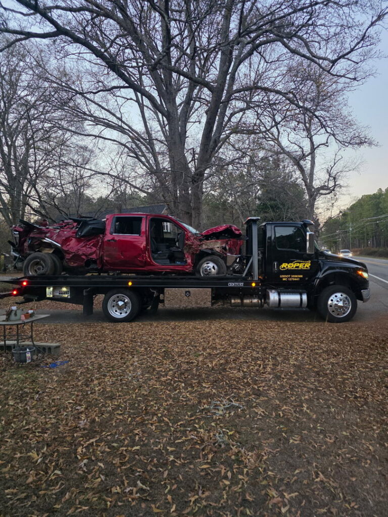 A Roper Automotive and Towing LLC flatbed tow truck hauling a heavily damaged red pickup truck in Greenville, SC.
