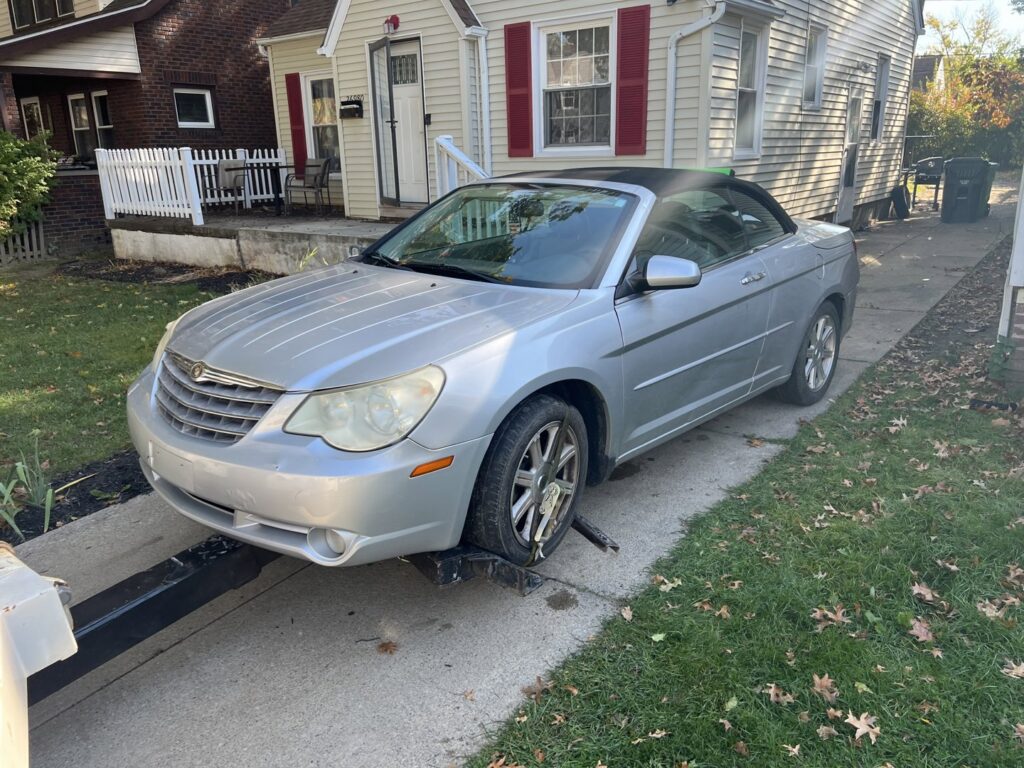 A tow truck from ALDM Management hooked to the front of a silver Chrysler Sebring convertible in a driveway in Cleveland, OH.