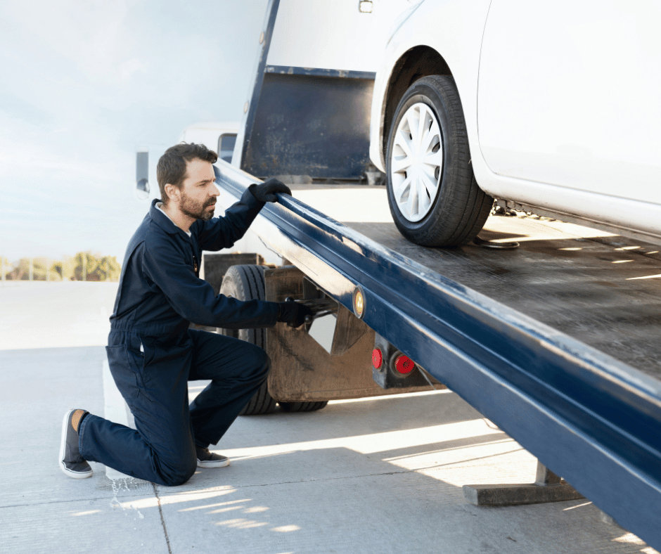 A tow truck operator in a blue jumpsuit securing a white car on a flatbed tow truck for Fort Benning Towing in Columbus, GA