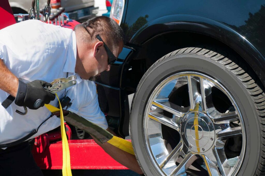A tow truck operator securing a black car's wheel with a strap on a flatbed for Spokane Towing Services in Spokane, WA.