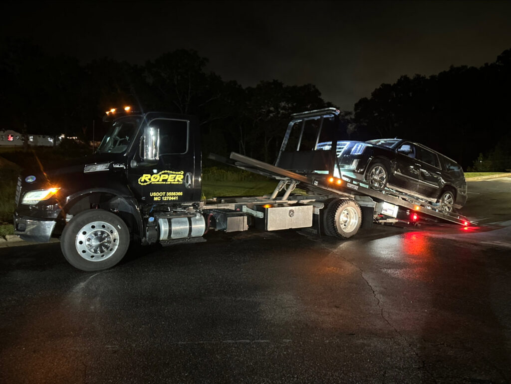 A Roper Automotive and Towing LLC flatbed tow truck recovering a black SUV at night in Greenville, SC.