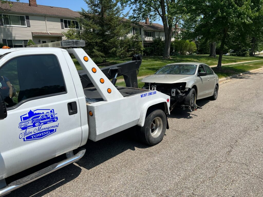 A white tow truck from ALDM Management towing a damaged silver car on a residential street in Cleveland, OH.