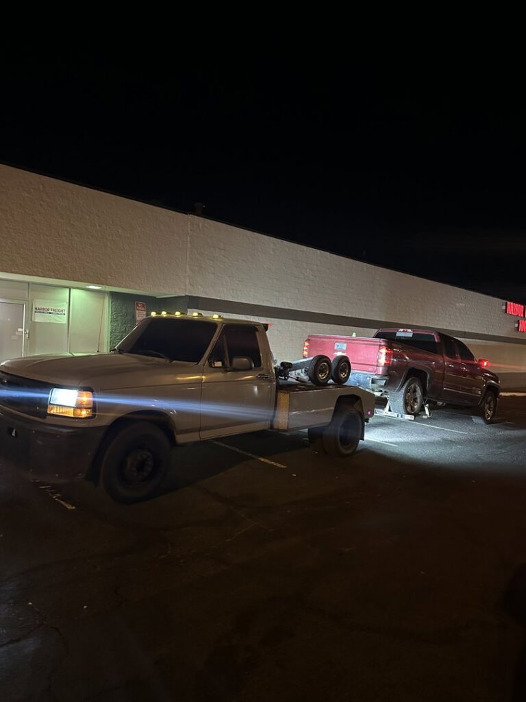 A tow truck from Henderson Towing & Recovery LLC towing a dark grey Ford Escape SUV in a parking lot in Denver, CO.