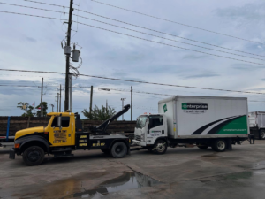 A yellow Houston Towing truck actively towing a white Enterprise box truck with a wheel-lift in Houston, TX.