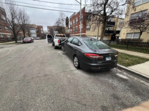A Trevi Towing llc truck towing a dark gray sedan with a dolly on a residential street in Chicago, IL.