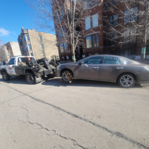 A tow truck from Soto Towing Inc. towing a brown sedan with a missing front wheel on a street in Chicago, IL.
