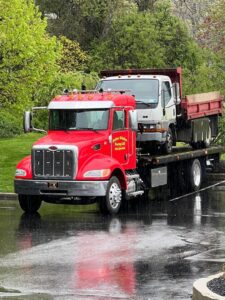 A red flatbed tow truck from Rubber Chicken Towing LLC towing a white dump truck in Charlotte, NC.