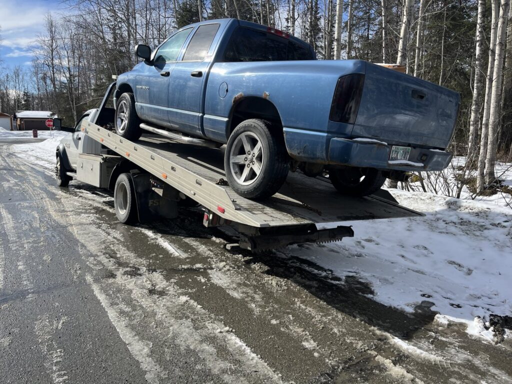 A Solid Choice Towing flatbed truck transporting a blue pickup truck on a winter road in Wasilla, AK.