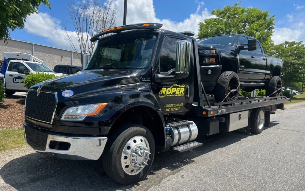 A Roper Automotive and Towing LLC flatbed tow truck transporting a black pickup truck in Greenville, SC.
