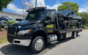 A Roper Automotive and Towing LLC flatbed tow truck transporting a black pickup truck in Greenville, SC.