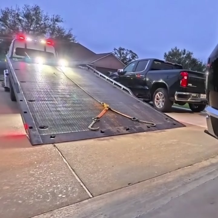 A tow truck with its bed lowered and lights on, ready to load a pickup truck at dusk, by Columbus Towing in Columbus, GA.
