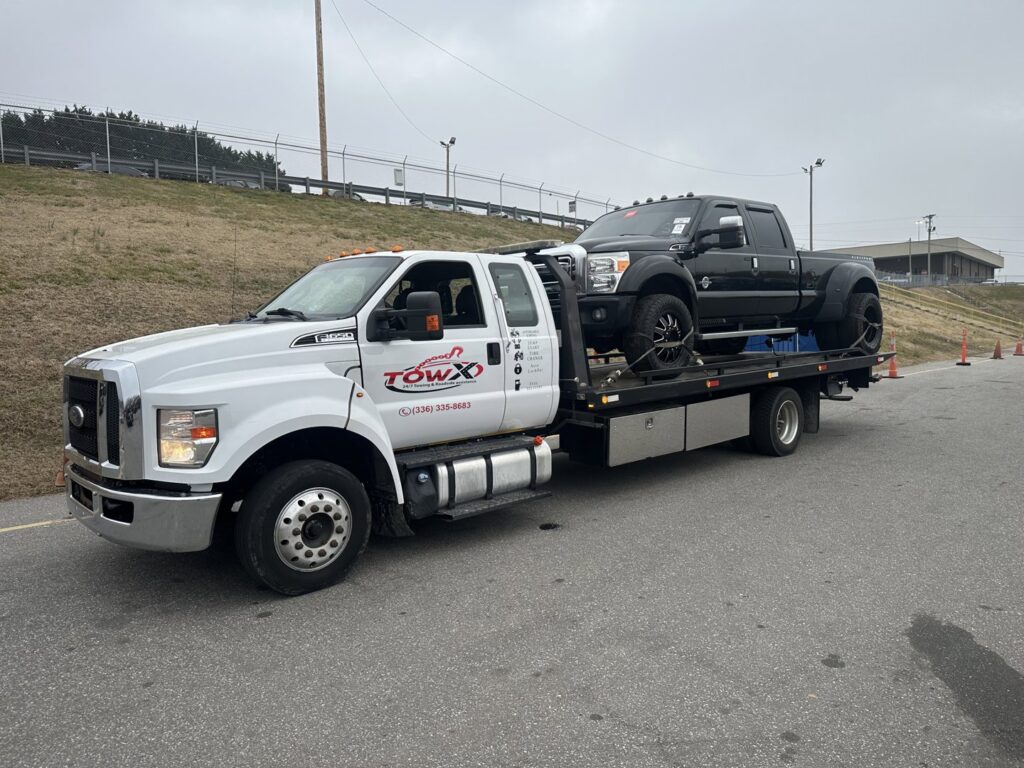A Tow X flatbed tow truck transporting a black pickup truck on a road in Greensboro, NC.