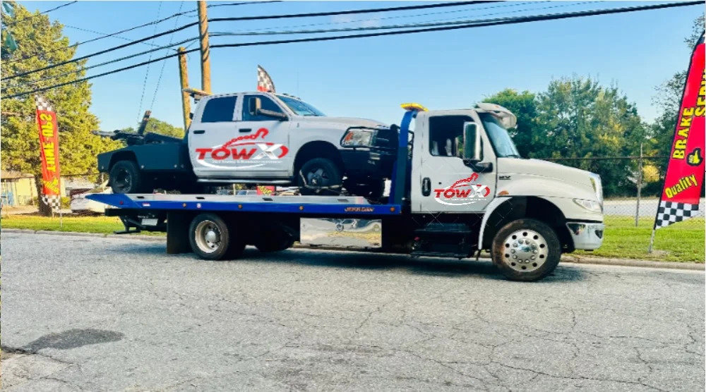 A Tow X flatbed tow truck transporting a white pickup truck with another Tow X wrecker behind it in Greensboro, NC.