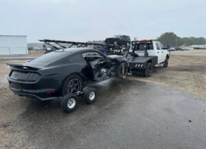 A Tow X truck towing a heavily damaged black Mustang with a dolly in Greensboro, NC.