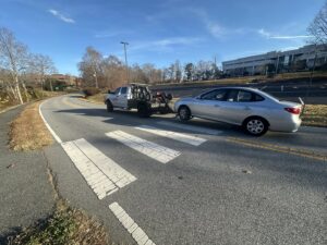 A Tow X truck towing a silver sedan with a dolly on a road in Greensboro, NC.