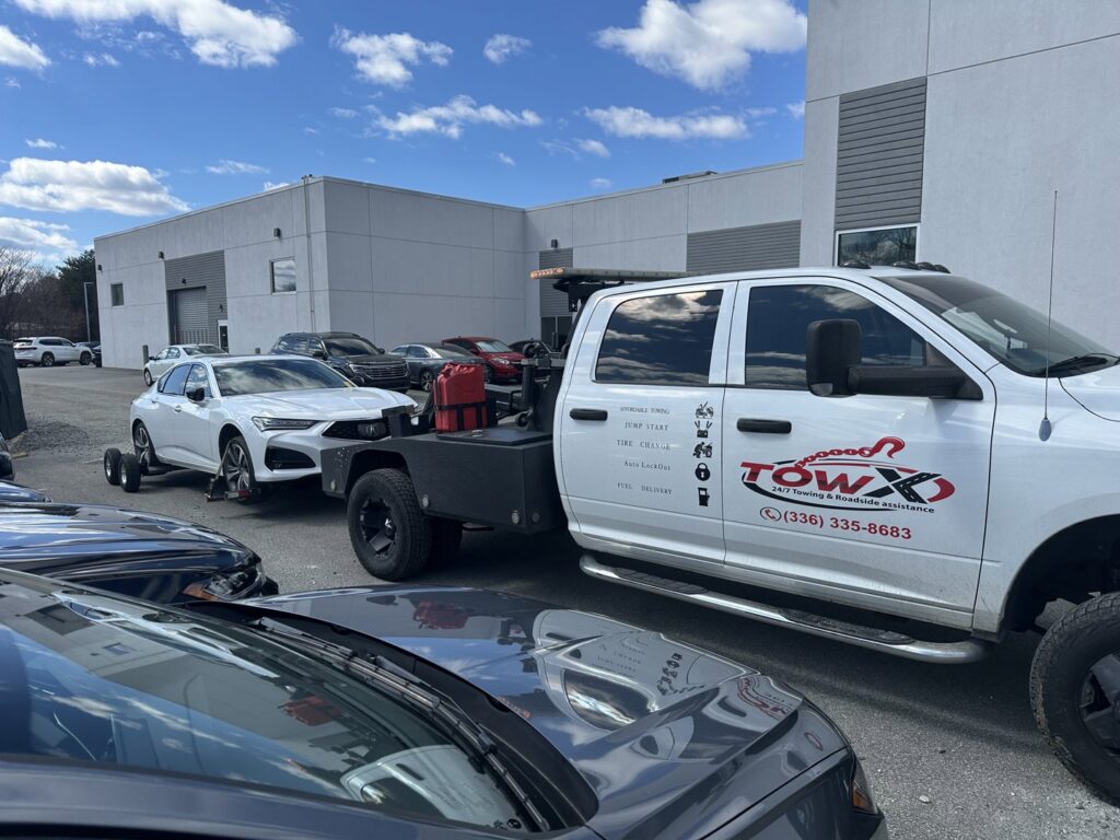 A Tow X truck towing a white sedan with a dolly in a parking lot in Greensboro, NC.