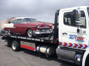 A Pacific Autow Center tow truck transporting a red and white classic car on its flatbed in San Diego, CA.