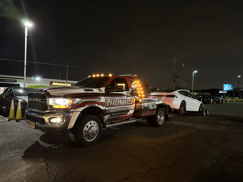 Thomas Towing & Recovery truck towing a white car on dollies at night in Newark, NJ.