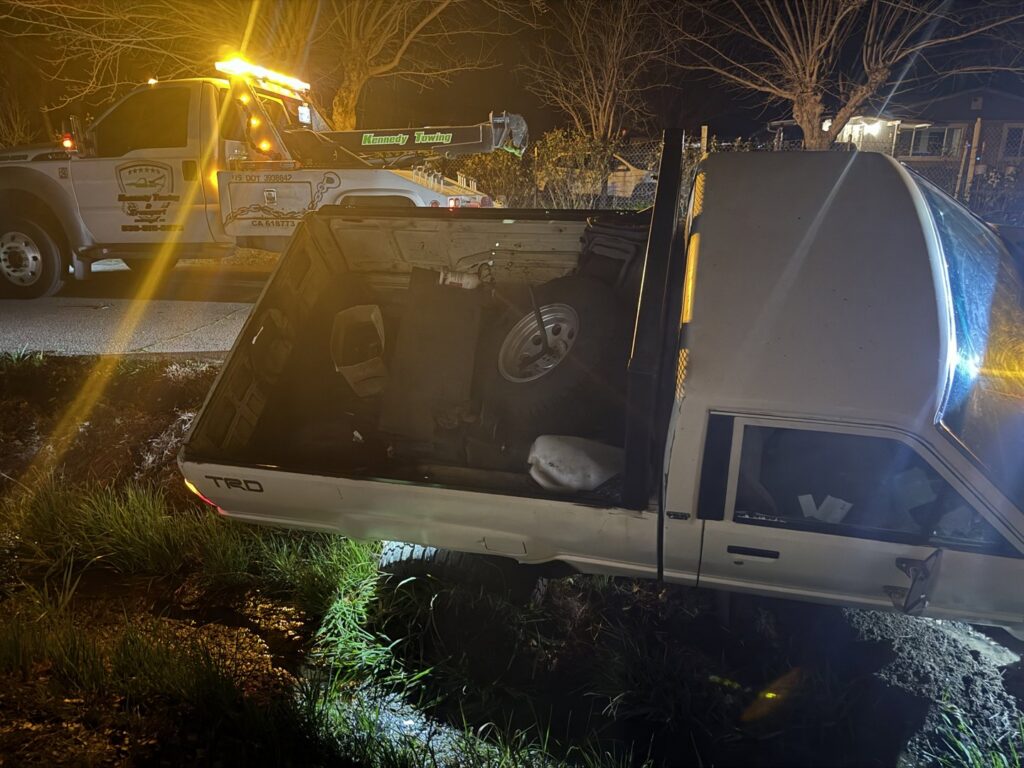 A white Toyota pickup truck stuck in a ditch at night, with a Kennedy Towing & Transport tow truck preparing for recovery in Anderson, CA.