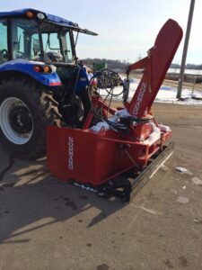 A blue tractor with a red Normand snow blower attachment ready for snow removal at Rochester Ground Lawn & Snow Services in Rochester, MN.