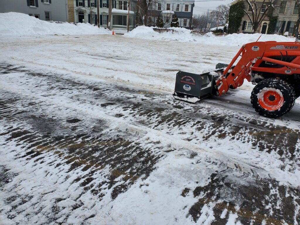 An orange tractor with a snow broom attachment clearing snow from a large parking lot by TPM Snow Removal in Schenectady, NY.