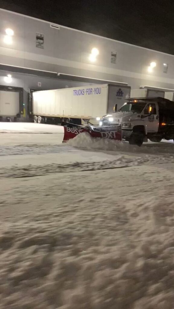A snow plow truck actively clearing snow from a commercial lot at night by Turf Pro Lawn Care & Snow Removal in Green Bay, WI.