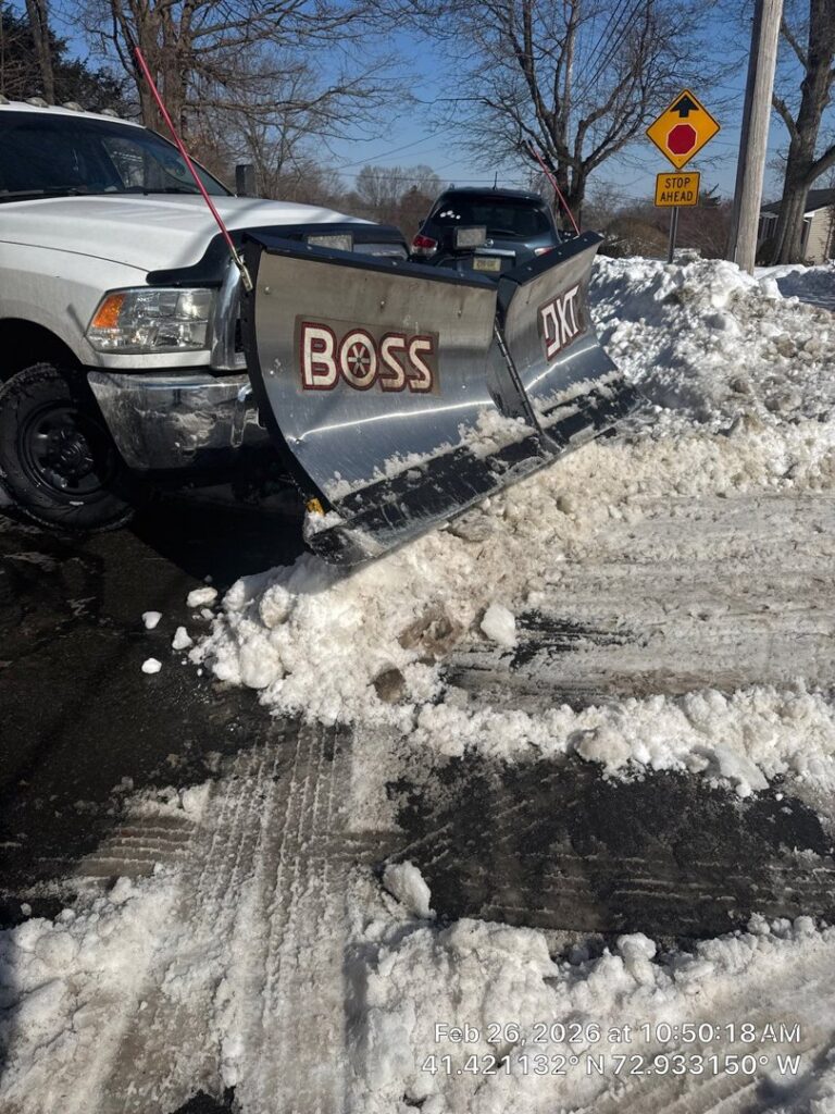 A white pickup truck with a BOSS DXT snow plow clearing snow from a street or large driveway by Diamond Image in New Haven, CT.