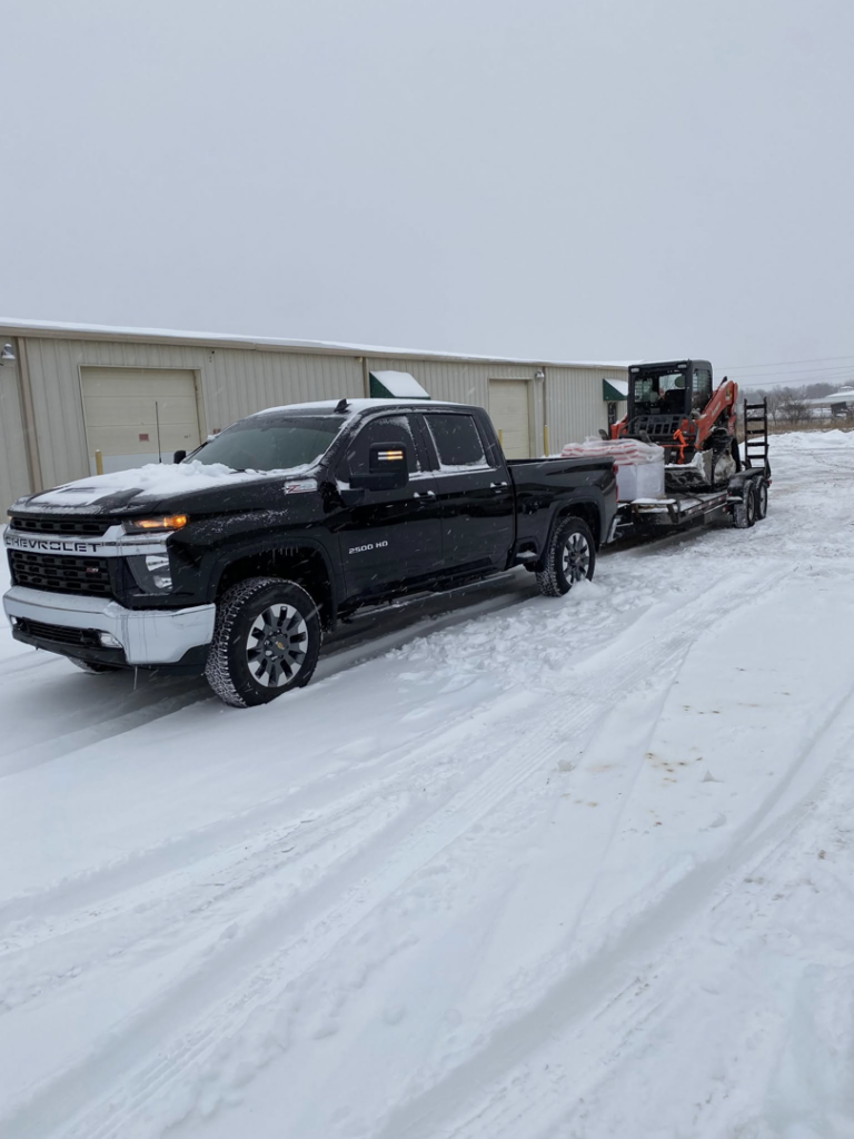 A black pickup truck towing a skid steer on a trailer through a snow-covered area for Creative Grounds in Springdale, AR