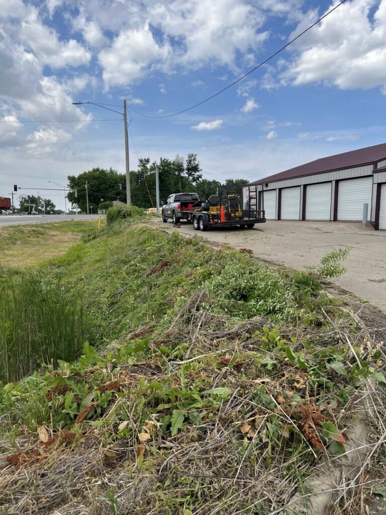 A truck and trailer with lawn care equipment parked next to an overgrown area, ready for clearing by Thompson Acres in Davenport, IA.