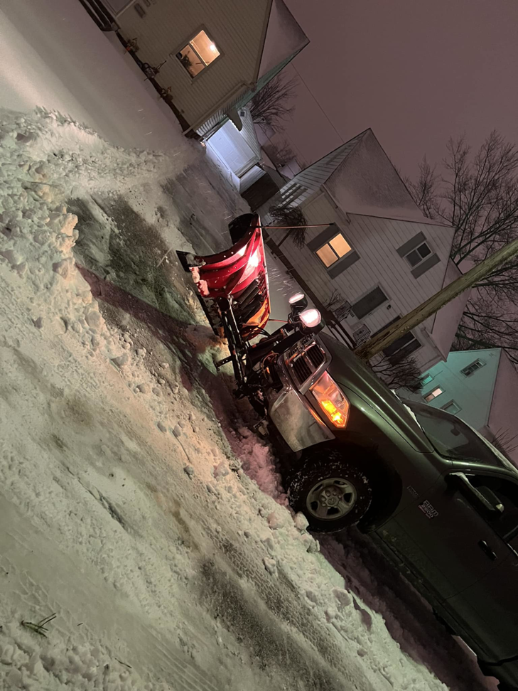A truck equipped with a snow plow clearing a residential driveway at night for PJs Full Service Contractors in Cleveland, OH.