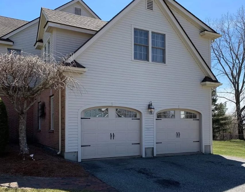 Two light-colored garage doors with decorative arched windows installed on a white house by NDI Garage Door in Plaistow, NH.