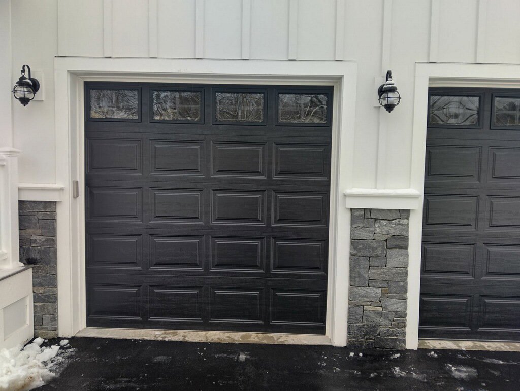 Two black panel garage doors with top windows and stone accents on a white house, installed by Steve Shumsky Unlimited Overhead Doors LLC in Hudson, NH