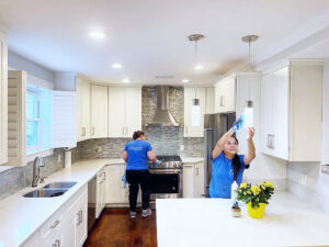 Two house cleaners from Supreme Home Cleaning, Inc wiping down surfaces and light fixtures in a bright kitchen in Boston, MA.