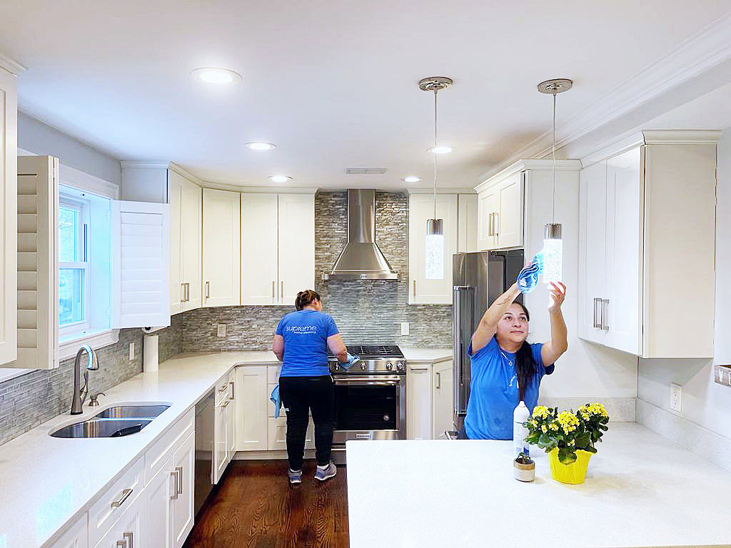 Two house cleaners from Supreme Home Cleaning, Inc wiping down surfaces and light fixtures in a bright kitchen in Boston, MA.